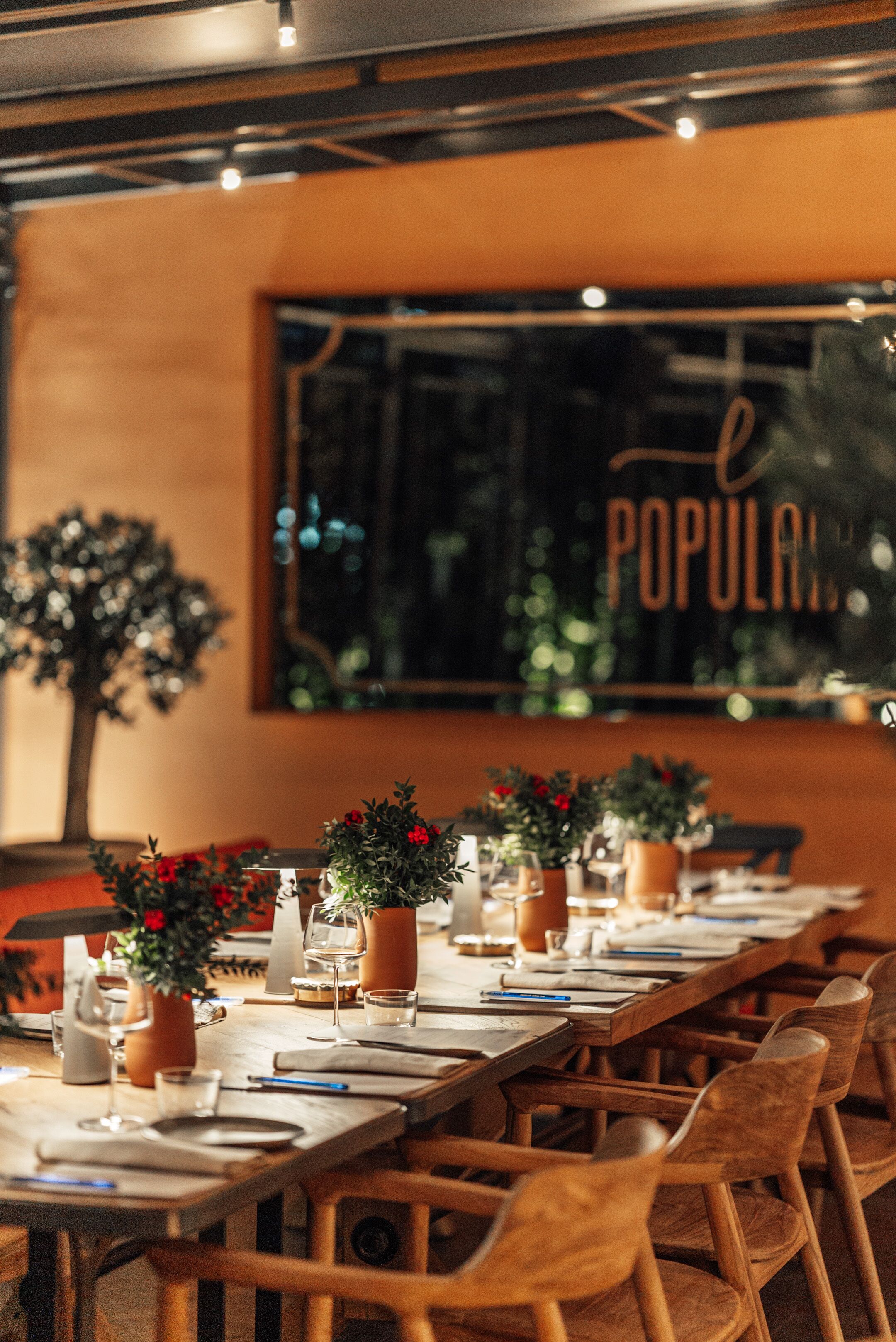 Un restaurante con una larga mesa de madera dispuesta para comer, plantas en macetas y un amplio ventanal con la palabra 'POPULAR' escrita.