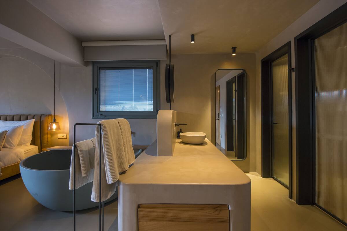 Ein modernes Schlafzimmer mit einer großen, runden Badewanne, einem Bett mit weißen Leinen, einem Nachttisch mit Lampe und einem Fenster mit Jalousien.