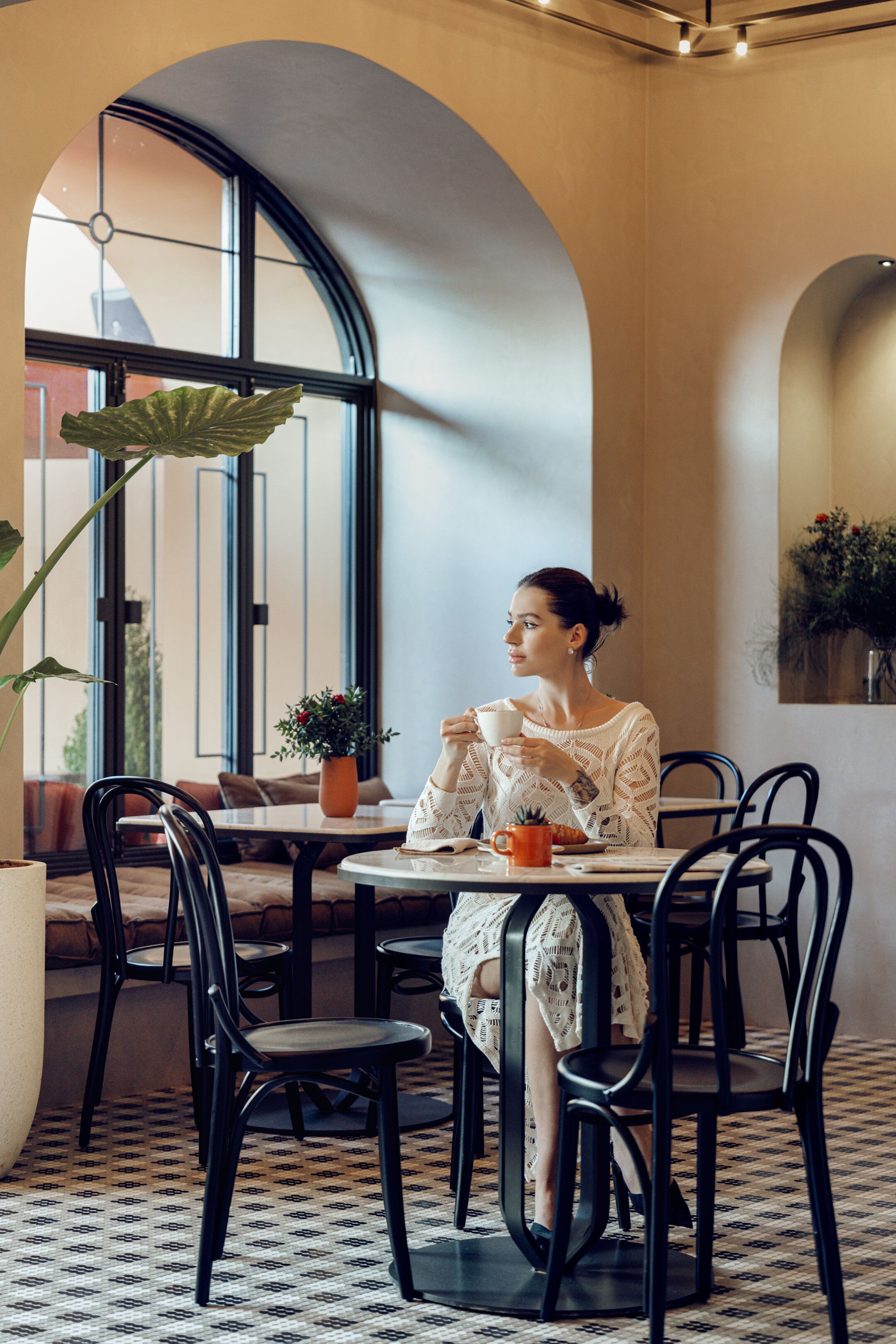 A woman sitting at a table in a cafe with a large window, black chairs, and a patterned floor.
