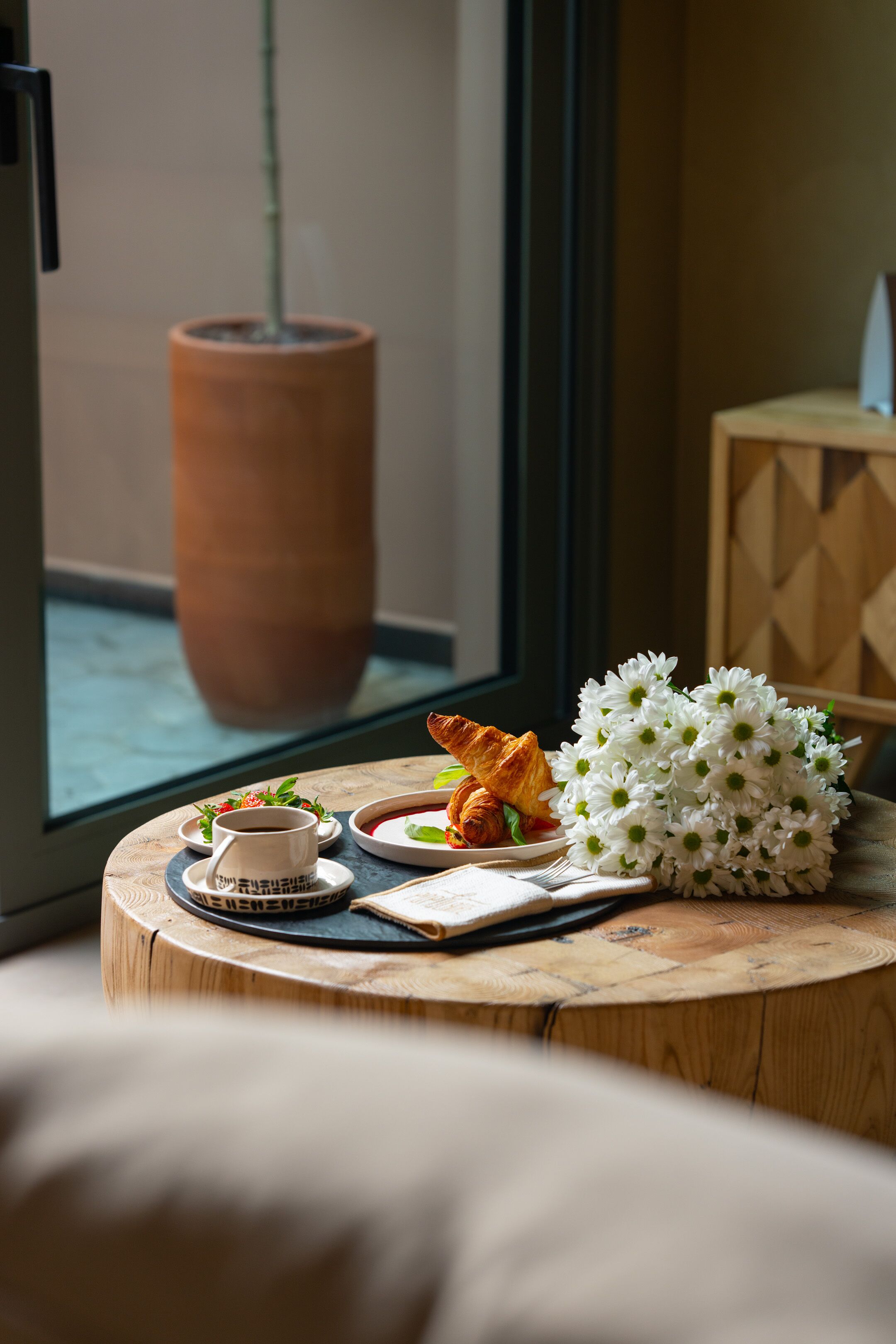 A breakfast setup with croissants, coffee, and flowers on a wooden table by a window.