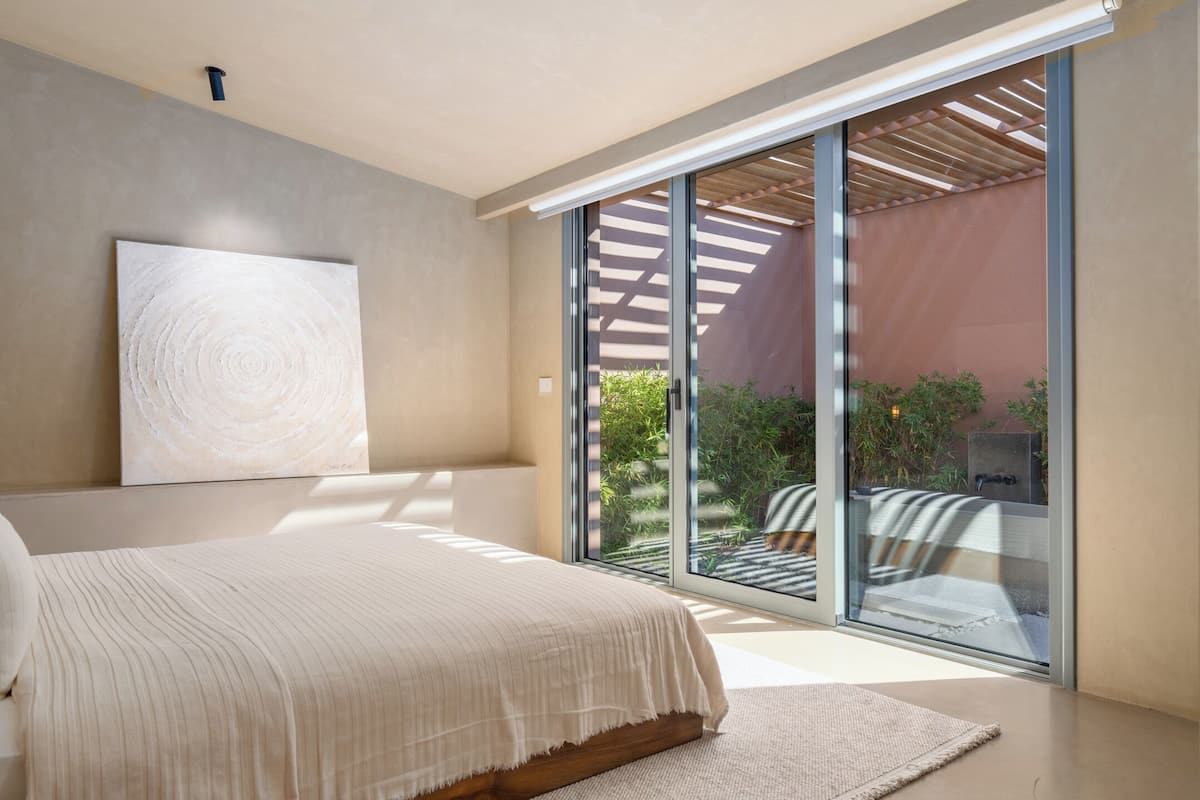 Ein Schlafzimmer mit einem großen Bett, einem an der Wand befestigten Kunstwerk und einer Glastür, die zu einem Außenbereich mit Terrasse und Pflanzen führt.