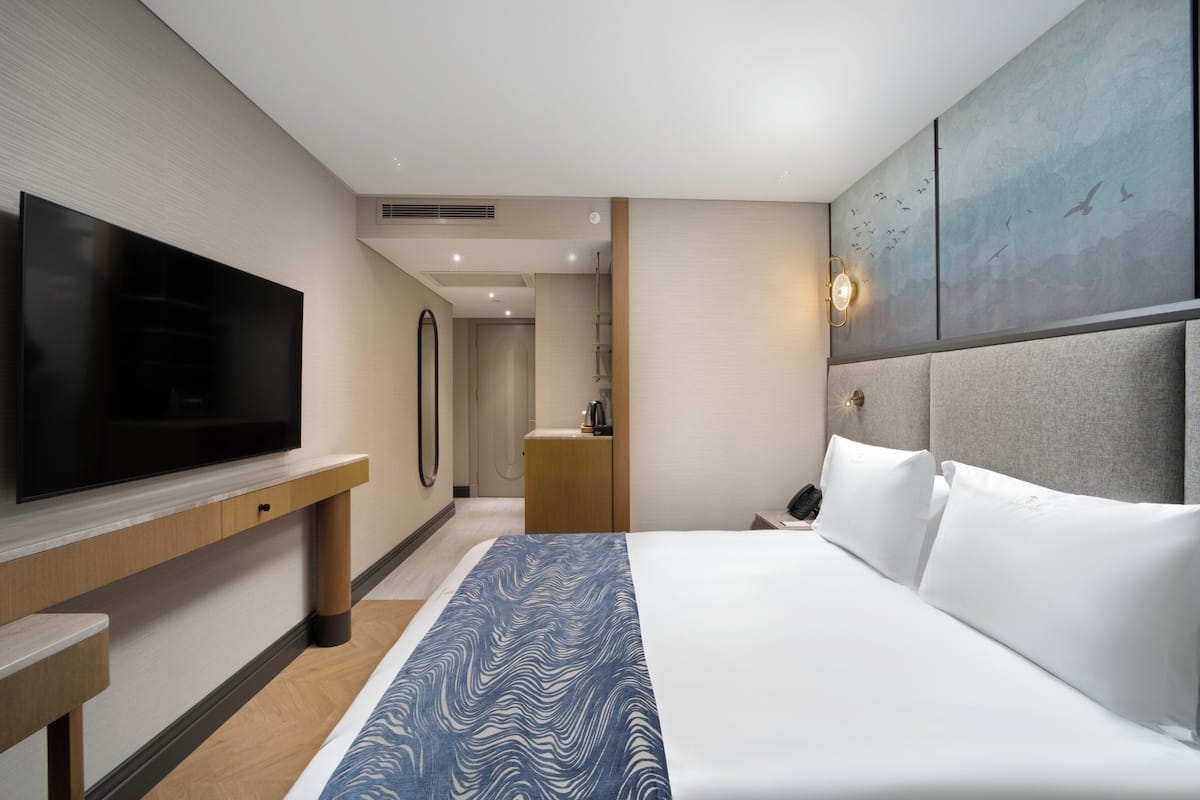 Habitació d'hotel amb un gran televisió de pantalla plana, un llit amb roba blanca, un estoraç de llit estampat de blau, una tauleta de nit de fusta i una obra d'art penjada a la paret.