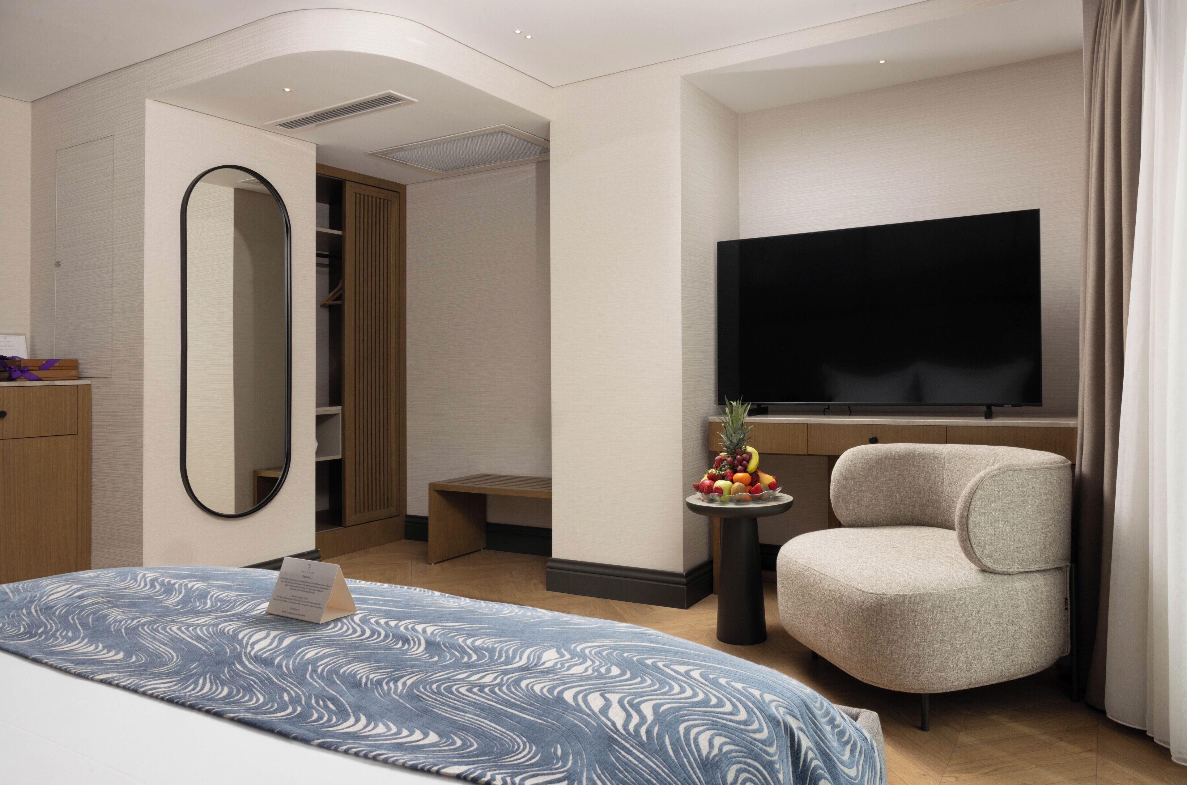 Une chambre d’hôtel avec un lit, une télévision, une chaise, une petite table avec des fruits et un miroir.