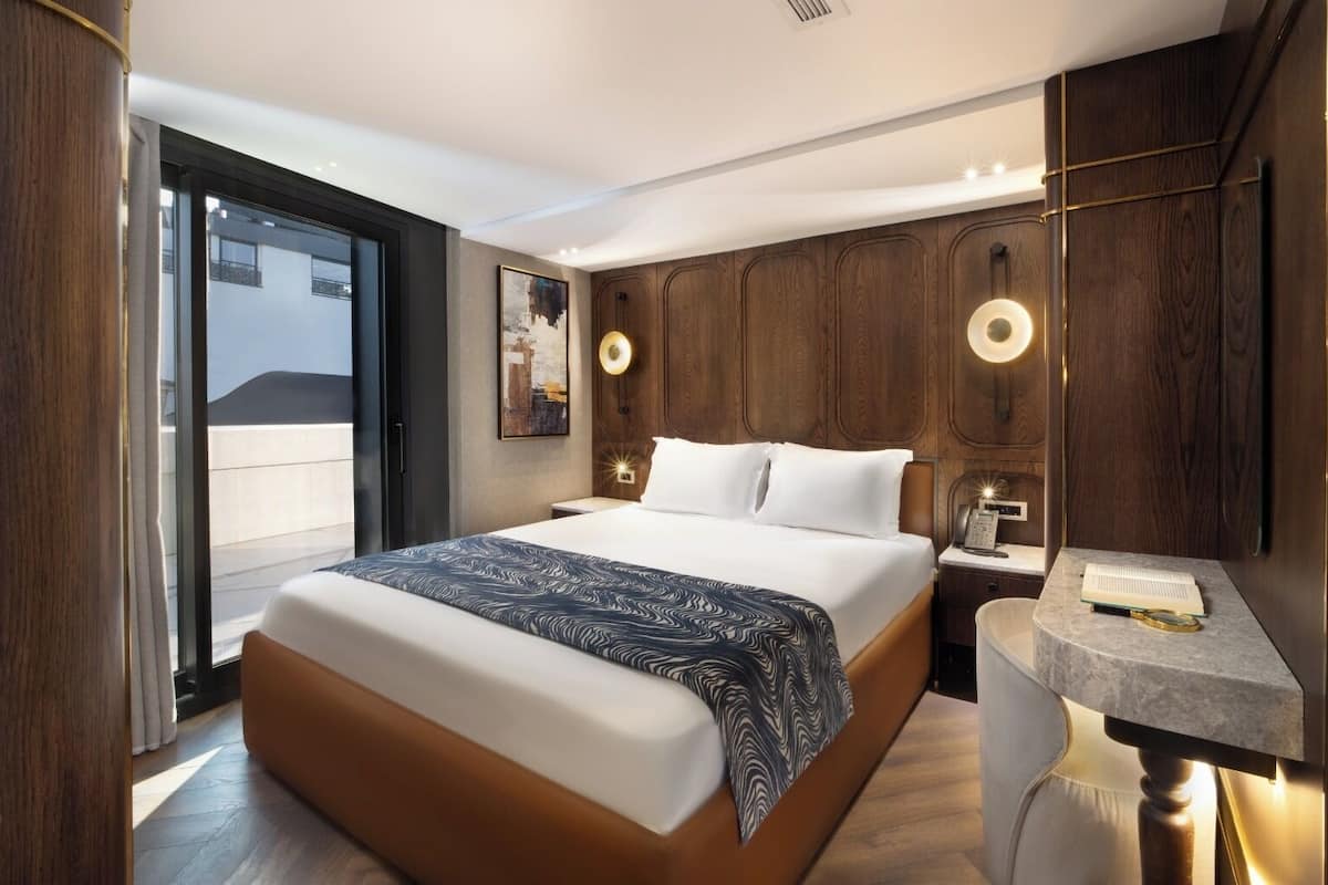 Una habitació d'hotel moderna amb un llit gran, panells de fusta, un escriptori amb una làmpada i una vista per una porta corredissa.