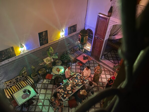 An indoor dining area with a tiled floor, multiple tables, and chairs. There are potted plants and decorative lighting.