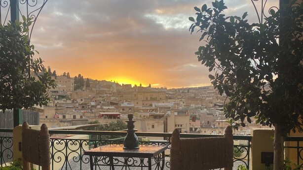 Un balcón con barandilla de hierro forjado, dos sillas de mimbre, una mesita y vistas a un paisaje urbano al atardecer.