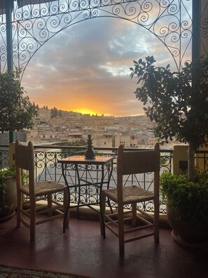 A balcony with a wrought iron railing, two wicker chairs, a small table, and a view of a cityscape during sunset.