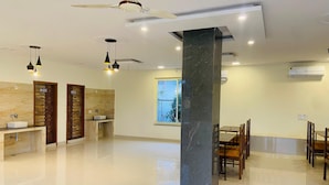 A modern hotel lobby with a central marble column, wooden wall panels, a dining area with wooden chairs, and a ceiling fan. - New Corbett Hotel and Resort (Ramnagar)