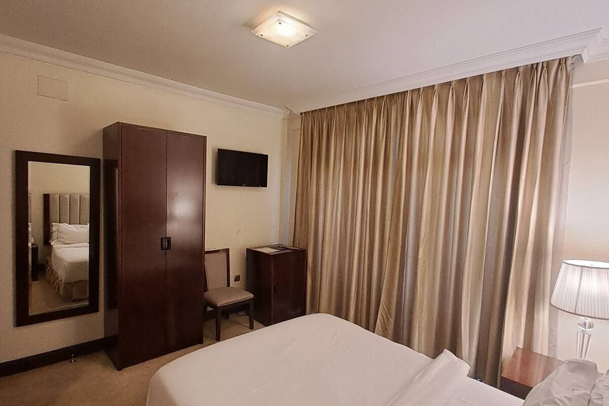 A hotel room with a large bed, a wardrobe, a desk, a chair, a television, and a window with curtains.