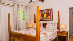 Un lit à baldaquin avec une couvre-lit à motifs floraux, deux tables de chevet avec des lampes, une œuvre d’art colorée accrochée au mur et une fenêtre avec des rideaux.