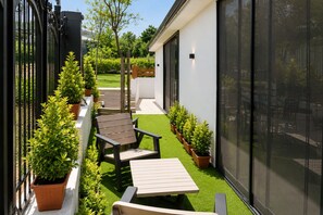 Une terrasse extérieure moderne, avec une table et des chaises en bois, entourée de plantes en pot et d’un bâtiment blanc doté de grandes fenêtres.