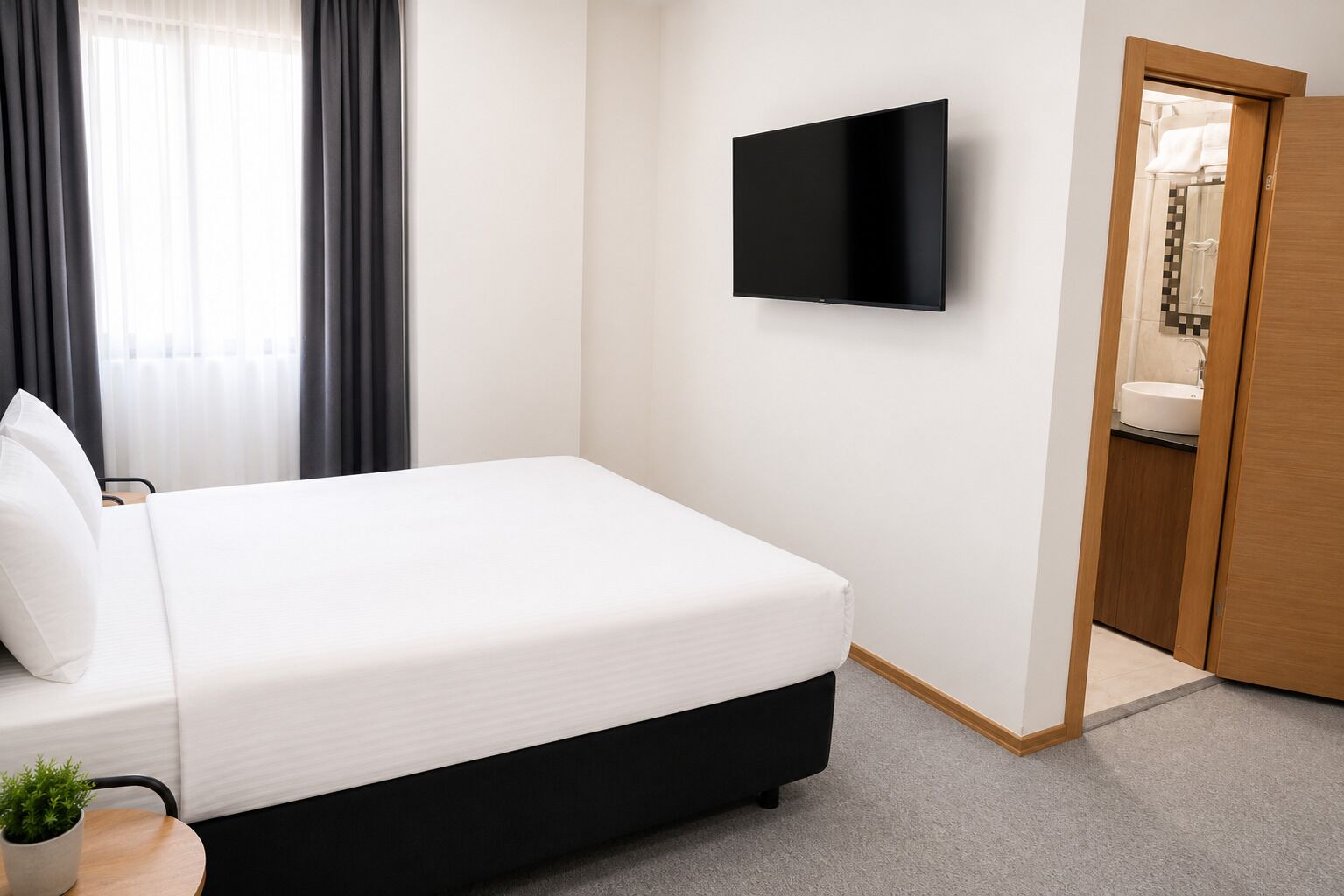 A hotel room with a bed, a flat-screen TV mounted on the wall, a wooden door, and a small table with a plant.