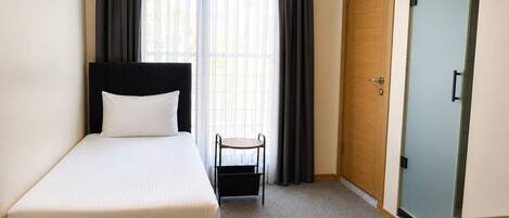 A single bed with a black headboard, a small table with a chair, and a wooden wardrobe in a room with a carpeted floor.