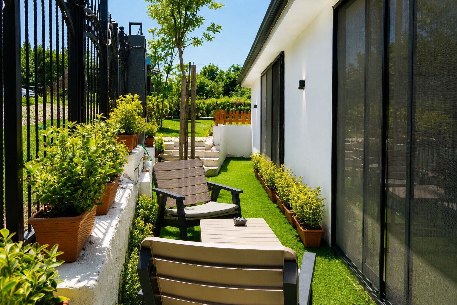 A patio with a chair and a table, surrounded by potted plants and a small lawn.