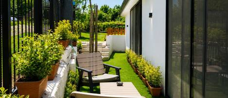 A patio with a chair and a table, surrounded by potted plants and a small lawn.