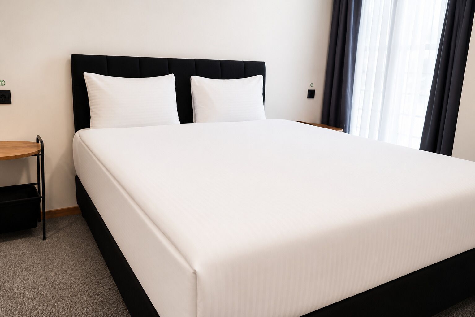 A neatly made bed with a black headboard and white bedding, a small wooden side table, and a power outlet on the wall.