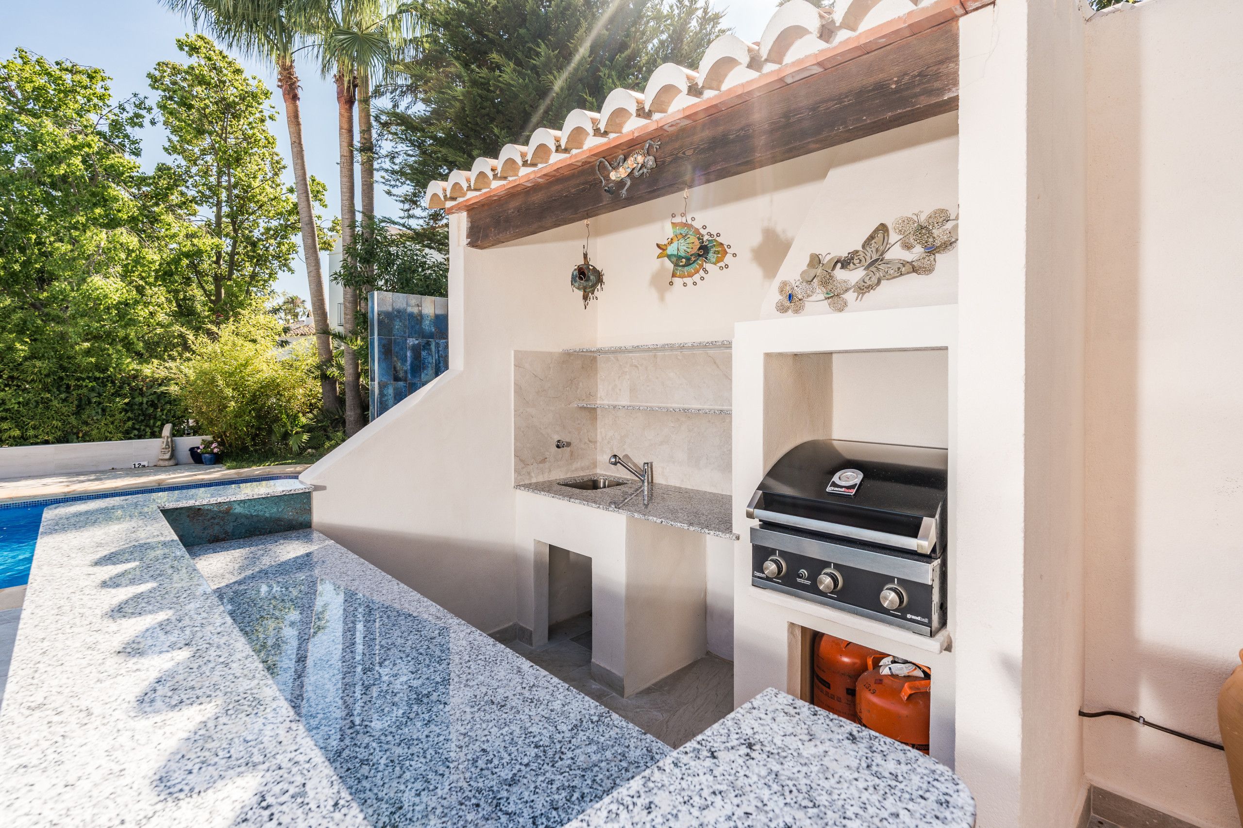 Cocina al aire libre junto a la piscina, con una parrilla empotrada, pileta y detalles decorativos en la pared.
