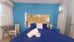 Una habitación con pared azul, cabecera de madera, cama con colcha azul, una silla blanca y una mesita.