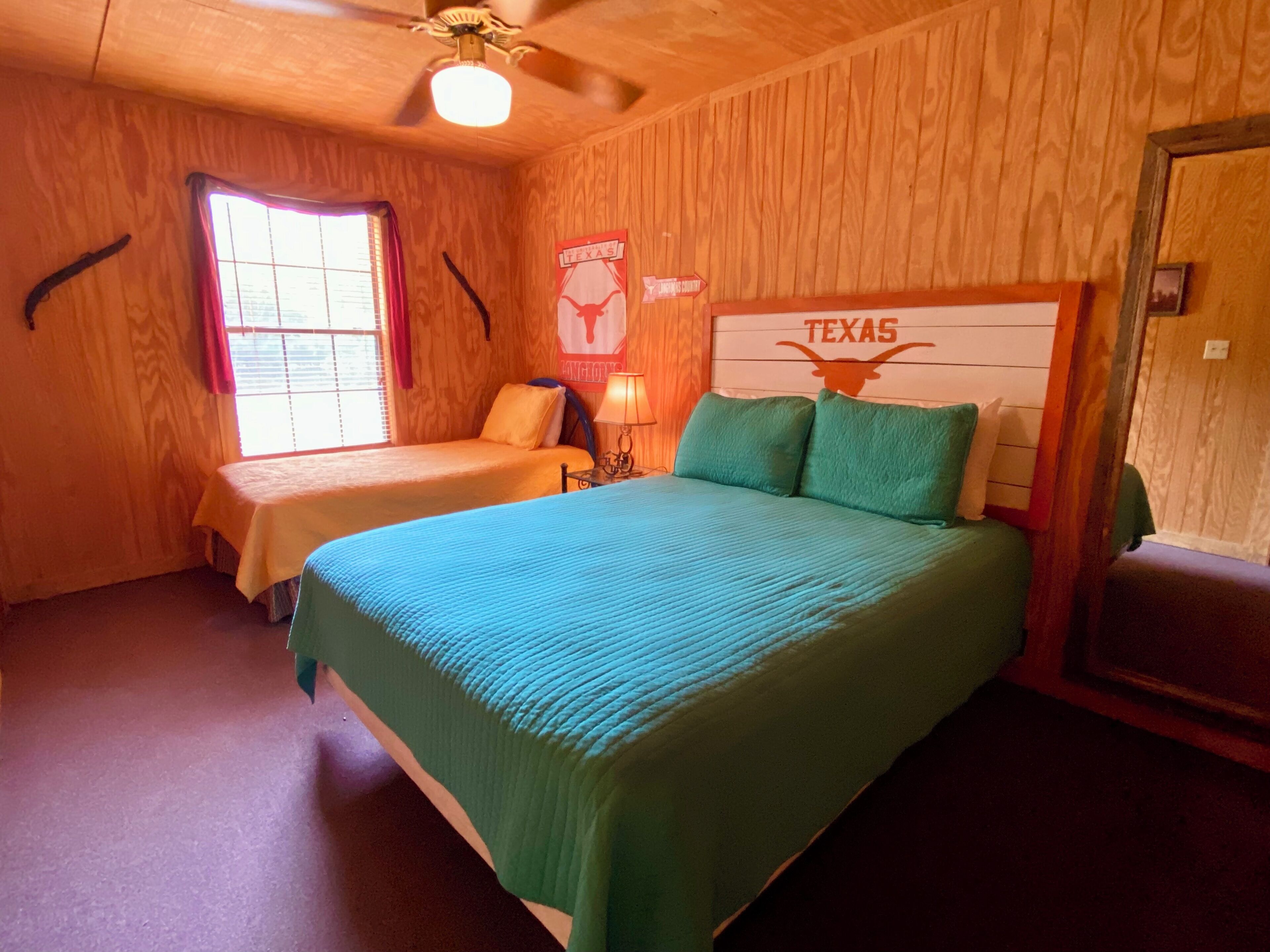 A room with two beds, wooden walls, a ceiling fan, and a window with blinds.