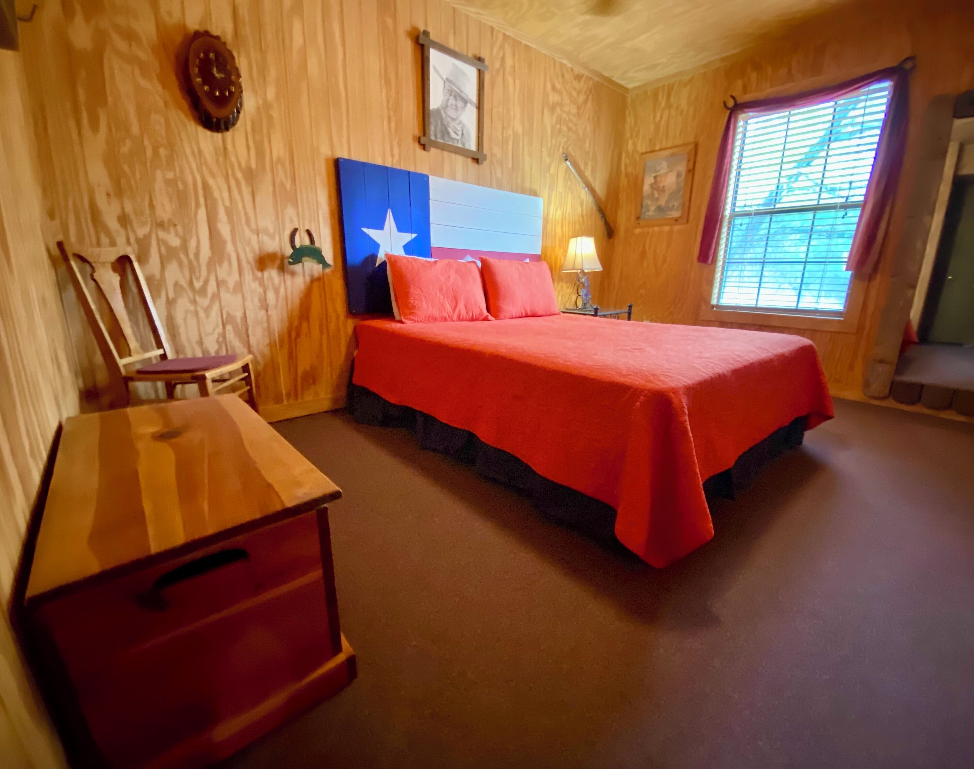 A room with a wooden wall, a bed with a red bedspread, a wooden dresser, a chair, and a window with blinds.