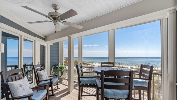 Une terrasse extérieure couverte, aménagée avec des meubles en bois et dotée de grandes fenêtres donnant sur la plage.