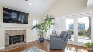 A spacious living room with a fireplace, a large TV mounted on the wall, a comfortable gray armchair, and a potted plant.