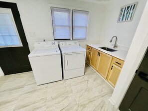 A compact laundry room with two washing machines, a sink, and wooden cabinets. - Vibrant 4BR Split-Level Home Near PTI Airport (Greensboro)