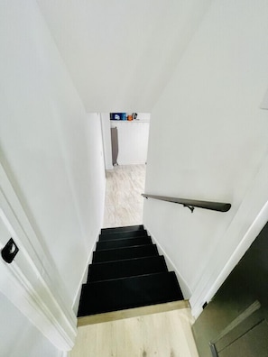 Eine Treppe führt zu einem Raum mit einer weißen Tür, Holzboden und einer kleinen Küchenzeile mit Kühlschrank und ein paar Gegenständen auf der Arbeitsfläche.