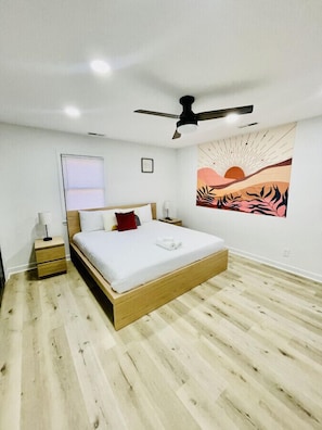 A modern bedroom with a wooden bed, white bedding, a ceiling fan, and a sunset painting on the wall.