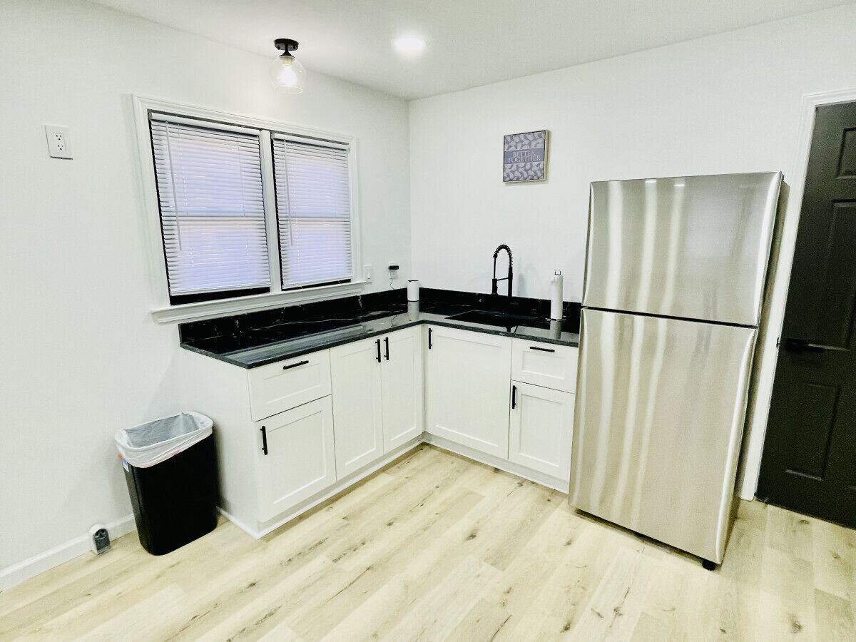 A compact kitchen with white cabinets, a stainless steel refrigerator, a black countertop, and a window with blinds.
