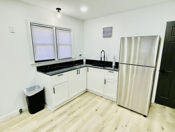 A compact kitchen with white cabinets, a stainless steel refrigerator, a black countertop, and a window with blinds.