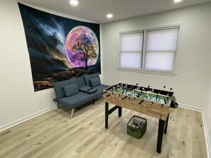 A room with a foosball table, a sofa, a window, and a wall tapestry.