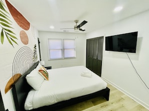 Un dormitorio con cama, ventilador de techo, televisor montado en la pared y ventana con persianas.