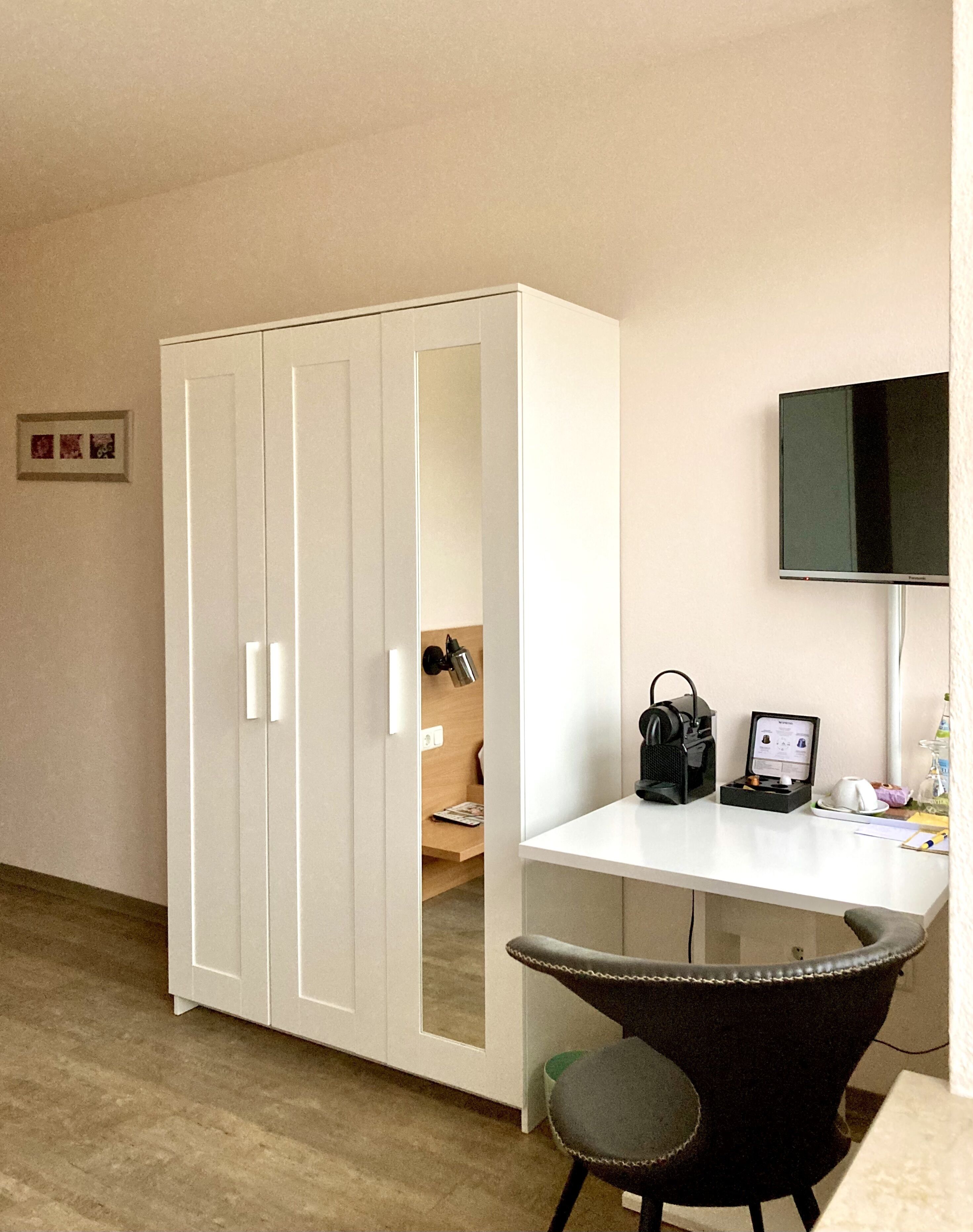 A room with a white wardrobe, a desk with a computer, a chair, and a TV mounted on the wall.