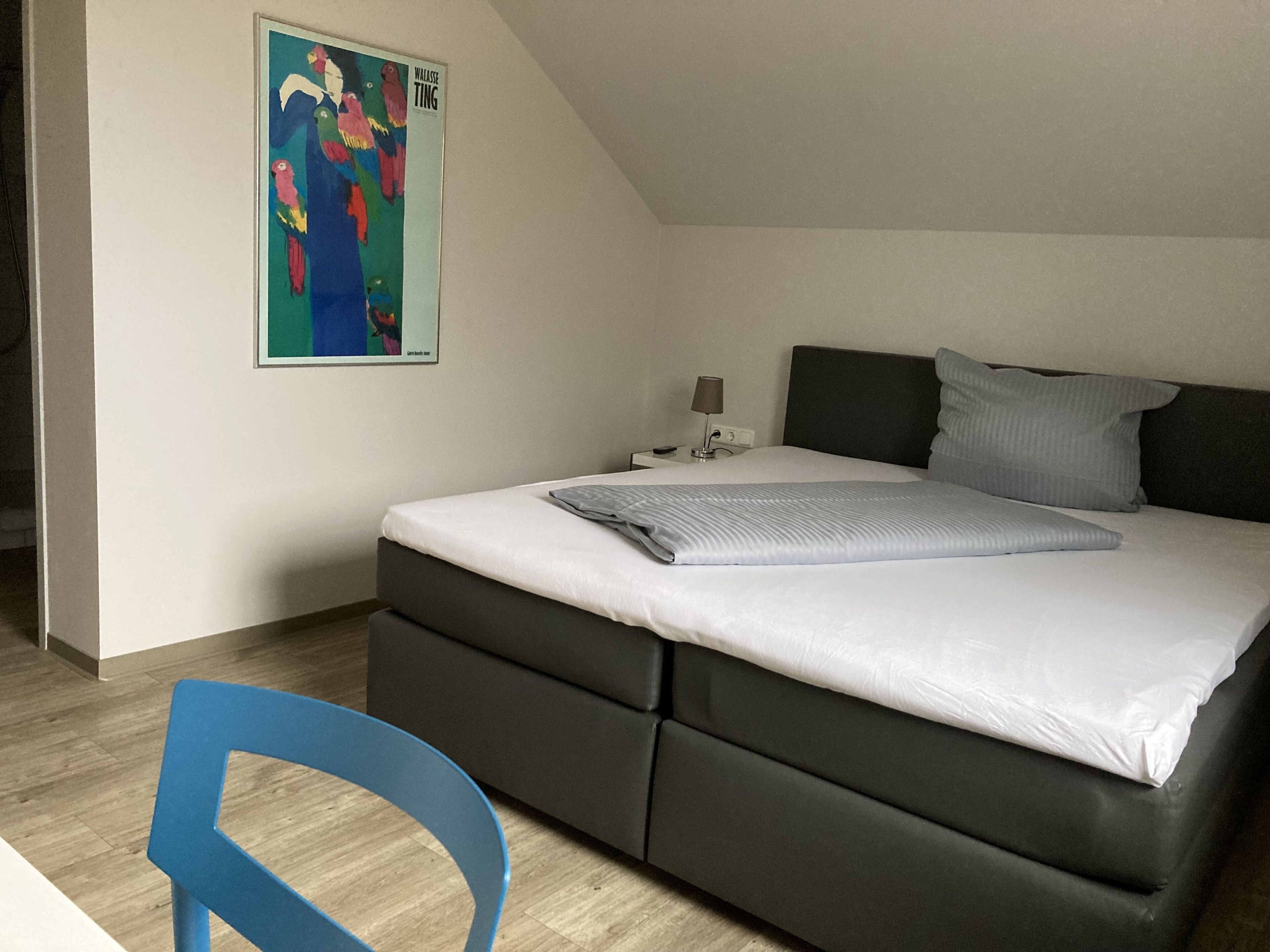 Un dormitorio con cama, un mesita de noche, una lámpara, una silla azul y un cuadro colorido en la pared.