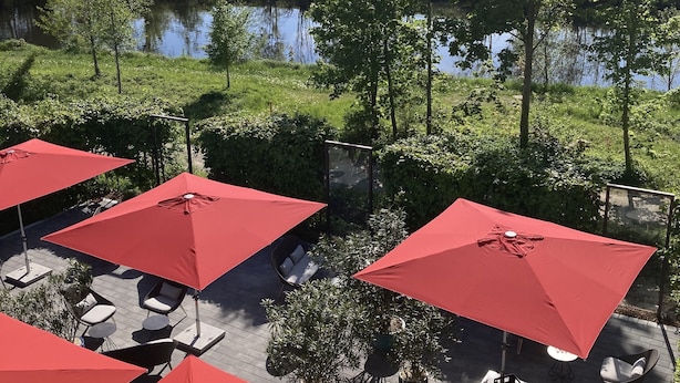 Un área con mesas y sillas al aire libre, con sombrillas rojas, y vista a un lago.