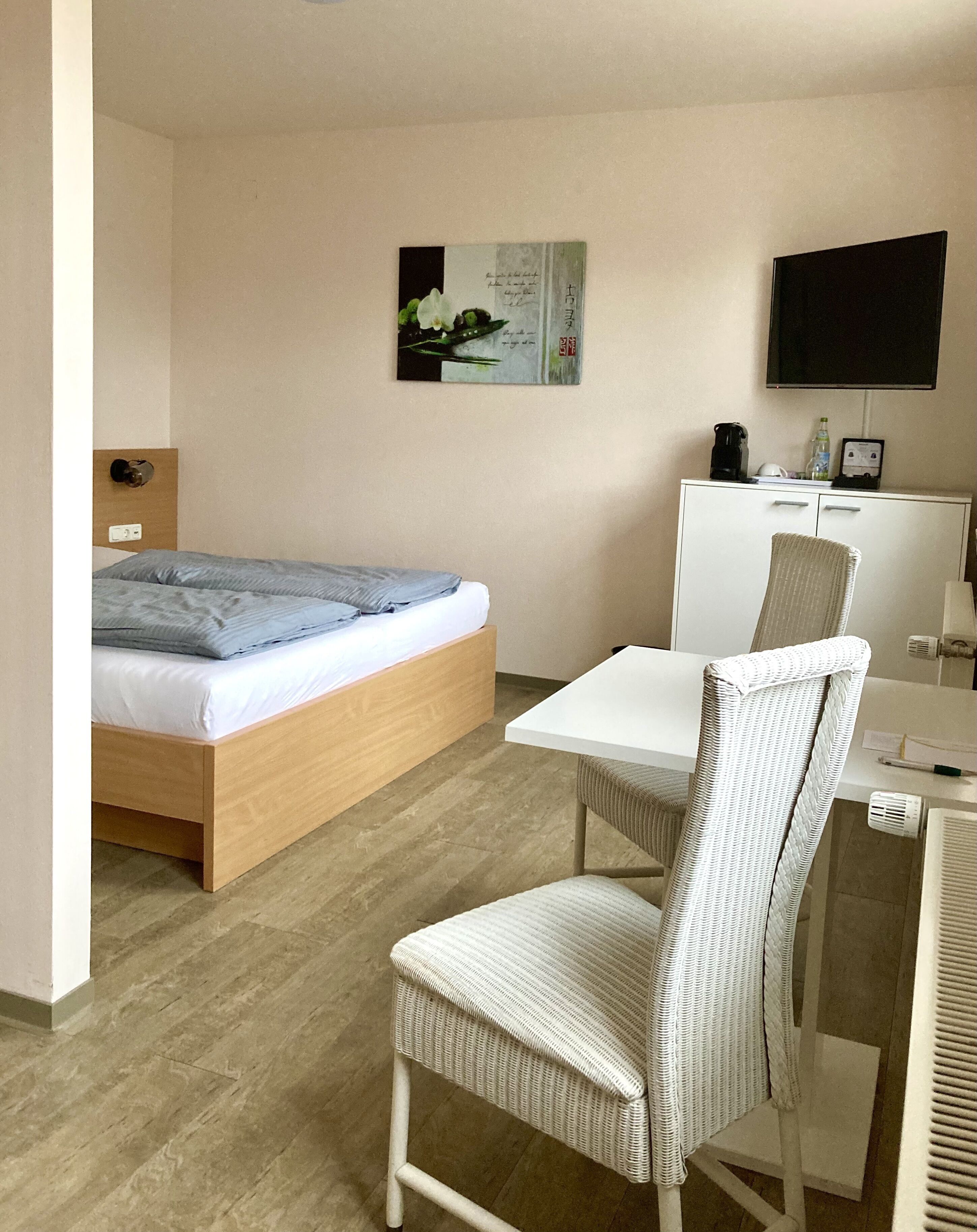 Camera d'albergo moderna con un letto di legno, una scrivania bianca, una poltrona in vimini, un televisore a schermo piatto e un armadio.