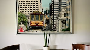 Una obra de arte enmarco que muestra un tranvía en una calle, una planta en maceta y dos sillas de madera.