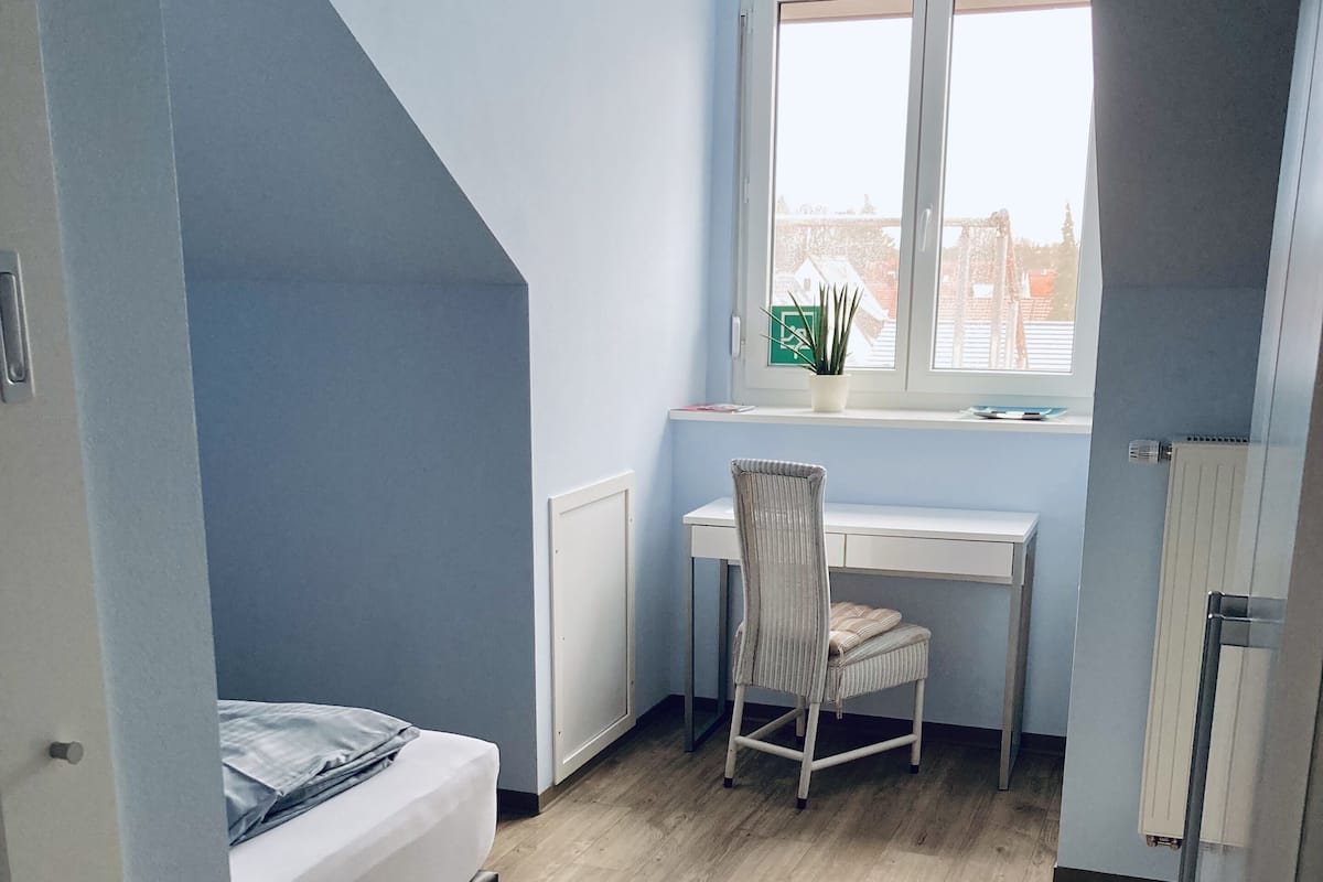 A compact room with a bed, a desk, and a chair. There is a window with a view of buildings outside.