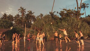 Eine Gruppe von Menschen spielt Beachvolleyball an einem Strand, im Hintergrund sind Palmen und Vegetation zu sehen.