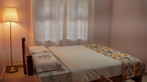 A neatly made bed with a floral-patterned bedspread and a white comforter, a bedside lamp, and a window with sheer curtains.