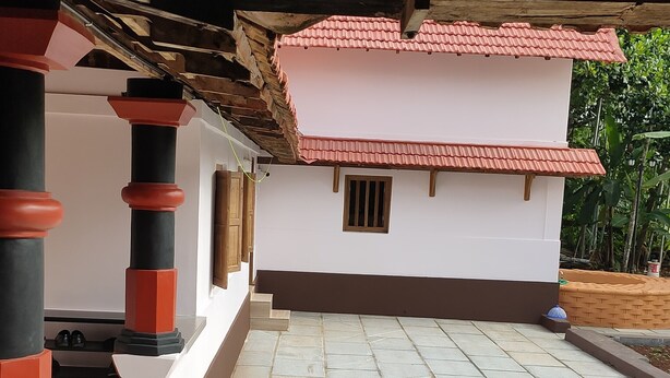 A covered outdoor area with a tiled floor, a white building with a red-tiled roof, and a wooden ceiling.