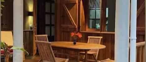 Een houten terras buiten, met een eettafel en stoelen, omgeven door witte zuilen en houten leuningen.