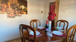 A dining room with a wooden table set for four, surrounded by chairs. There is a painting of fruits and vegetables on the wall, and a decorative vase with red flowers on the table. - The Gold- Home Away From Home (Manchester)