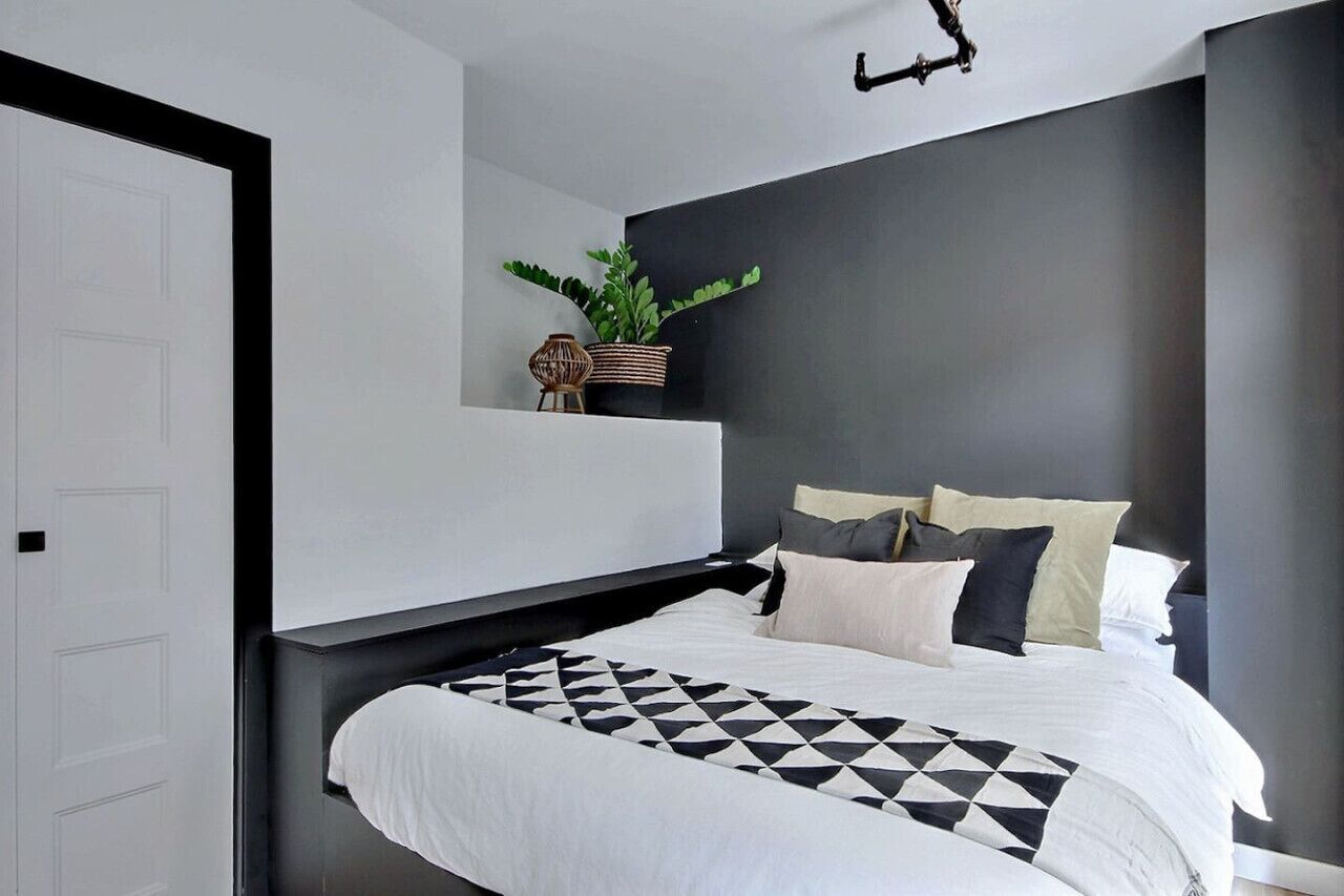 A modern bedroom with a bed, a nightstand, a potted plant, and a wall-mounted light fixture.