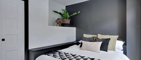 A modern bedroom with a bed, a nightstand, a potted plant, and a wall-mounted light fixture.