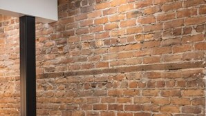 A hotel room with a brick wall, a black headboard, a white bed, a black nightstand, a white lamp, and a gray pillow. - Château Deluxe / / Vieux-Québec, Énorme Maison (Québec)
