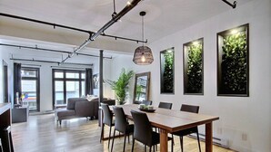 Uma zona de refeições moderna com mesa e cadeiras de madeira, janelas amplas e um lustre.