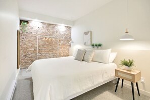 A bedroom with a brick wall, a bed with white bedding, a bedside table with a plant, and a hanging light fixture.