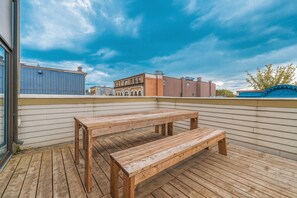 Eine Holzterrasse mit Tisch und Bank, ein blaues Gebäude mit der Aufschrift '1905' und ein klarer Himmel.